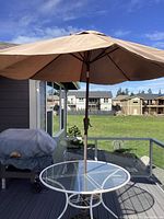 Round metal framed patio table with clear glass top on outdoor deck with beige umbrella open.