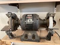 Photo of the bench grinder showing the entire unit with both grinding wheels, metal tool rests, and base plate on a wooden surface.