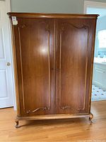 Front view of the wooden armoire highlighting the two large doors with carved decorative panels, the rounded top edge, and the sturdy cabriole legs.