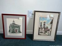 Two framed and matted architectural paintings leaning against a wall, showing a colonial style building and a larger ornate building with towers and domes.