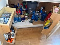 Wide view of under-sink cabinet with multiple cleaning products mostly in blue, white, and brown plastic bottles on shelves and door organizers.