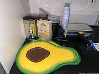 Coffee grinder, coffee filters box, two avocado-shaped placemats, pizzelle maker, and water bottle on kitchen counter
