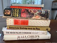Stack of six vintage cookbooks including Betty Crocker's Picture Cook Book, Julia Child's Menu Cookbook and German and French titles, showing wear on spines and edges.