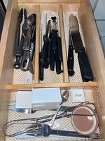 Drawer containing assorted kitchen knives, peeler, serving spoons, tongs, and other utensil tools with black handles.