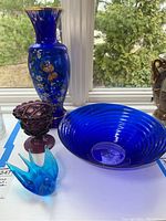 Photo of the four items: large blue glass bowl, blue glass bird figurine, purple textured glass bowl, and tall painted blue vase