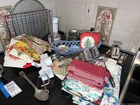 Photo showing assorted kitchen linens, a hand-crank egg beater, metal graters, a ceramic basket, and a Christmas-themed ceramic holder on a surface in front of tiled wall.
