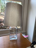 Full view of the glass table lamp with beige fabric lampshade on wooden surface near window.
