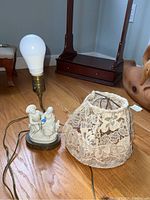 Figurine lamp with lace lamp shade removed and displayed beside it, showing the lamp base with figures and modest lamp bulb attached.