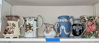 Full view of six pitchers on a shelf showing different designs and colors, some floral, some classical reliefs.