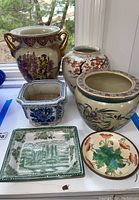 Photo showing the six ceramic items including various planters and decorative plates with Asian style and floral designs on a white table near a window.