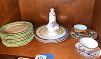 Shelf display featuring a stack of green floral dessert plates, a white condiment shaker with purple floral pattern, and two Japanese tea cups with matching saucers featuring colorful designs.