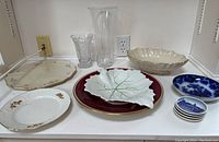 Multiple porcelain and glass items including Lenox bowl and platter, Limoges tray, various plates, and glass vases arranged on a shelf.