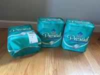 Three packs of Prevail absorbent protection pads in blue-green packaging on hardwood floor, each containing 15 pads.
