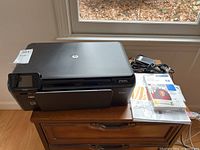 HP Photosmart D110a printer with power cable, manuals, and photo paper on wooden floor by window.