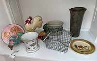 Shelf display of Royal Doulton plate on stand, ceramic pot, rooster figurines, pottery containers, metal vase, and wire baskets