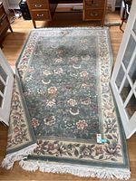 Full view of the floral green rectangular rug fully spread on a wood floor with one corner partially folded to show thickness and underside.