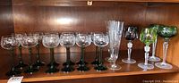 Full view of stemware collection on wooden shelf including green stem glasses, clear glass flutes with etching, and colored glasses