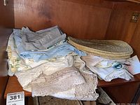 Stack of assorted cloth napkins, lace-edged tablecloth, various fabric tablecloths and woven gold placemats on wooden furniture shelf.