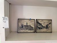 Two framed German clay tiles with black and white illustrations of Wangerin scenes on a white background placed on a shelf.