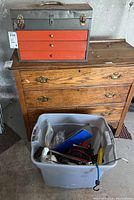 Metal vintage tool box with three orange drawers showing wear and rust, placed on a wooden dresser next to a plastic bin of tools.