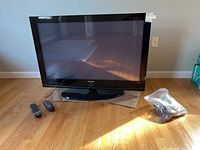 Samsung 42 inch standing TV on floor with two remotes and sealed accessories bag nearby.
