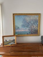 Two paintings displayed on a wooden surface against a white wall: a smaller signed painting by Hans with a gold-tone frame and a larger Monet-style tree landscape painting in an ornate gold frame.