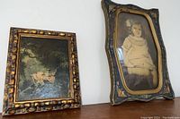 Two framed vintage items displayed side by side on a wooden surface. Left: vintage print painting in gold-accented carved frame. Right: vintage child's photo in ornate floral decorative frame with gold trim.