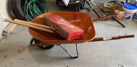Wide view of orange metal wheelbarrow with wooden handles holding a red Milwaukee Super Sawzall case and 2 wooden-handled axes