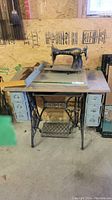 Full view of antique Singer sewing machine mounted on wooden table with drawers and cast iron base in workshop environment.