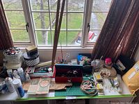 Wide view of sewing and craft tools arranged on a table by a window showing embroidery hoops, containers, spray cans, and boxes
