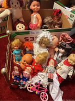 Photo of assortment of various vintage dolls including Raggedy Ann dolls, Kewpie doll, baby dolls, ballerina doll in costume, brass doll bed, and small pink toy bicycle.