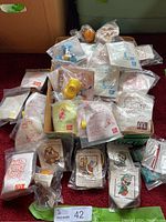 Various unopened McDonald's Happy Meal toys in original sealed plastic bags arranged in a box and surrounding area, including Joe's Mooty Boo's and Peanuts collections.