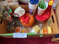 View of multiple McDonald's collectible items inside a box including glass tumblers, pins, plastic containers shaped as red shoes on orange base, a white Ronald McDonald mug, and souvenir plastic cups.