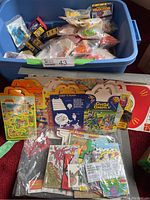 Wide view showing large blue plastic bin filled with sealed McDonald's Happy Meal toys in bags, with numerous vintage Happy Meal boxes and activity sheets arranged on the floor.
