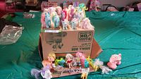 Photo of various My Little Pony toys arranged on a cardboard box on a table with a green cover. Shows an assortment of ponies in pastel colors with manes of different colors.