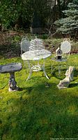 Outdoor setting with white metal bistro table and two chairs, birdhouse decor on table and chair, stone birdbath and stone rabbit statue on grass.