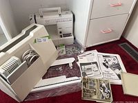 Wide view showing Pfaff Hobbymatic 927 sewing machine with attached power cord, white plastic carrying case, manuals, and a box of assorted sewing machine parts on a red carpet.