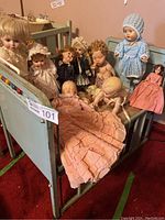 Front view photo of the blue metal doll crib filled with 10 vintage dolls, showing detailed faces and clothing of dolls and crib construction.
