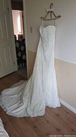 Full length side and front view of the white wedding gown hung on a hanger showing long train and fitted silhouette.