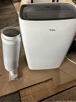 TCL portable air conditioner main unit with white flexible exhaust hose and window vent adapter placed on floor
