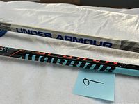 Close-up photo of two lacrosse shafts showing 'Under Armour' and 'Warrior' branding.