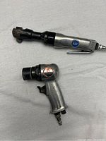 Two pneumatic air tools on a grey cloth: one air ratchet wrench with a silver body and a black drive end, labeled '1/2 inch drive'; one pneumatic air sander with pistol grip and sanding head, labeled with safety warning.