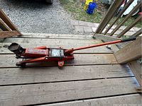 Orange hydraulic car floor jack positioned on wooden decking with vehicle in background, showing its side profile and lifting parts.