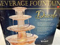 Full box image showing the Rival Dazzle Beverage Fountain and marketing points including bubbles, colored lights, serving up to 2.5 gallons, and dishwasher safe.