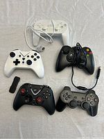 Five gaming controllers arranged on a white fabric background, featuring white and black color schemes, wired and wireless types, varying button layouts.