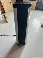 Front view of Pelonis tower heater standing upright, showing black grille and white body with Pelonis branding at base.