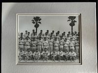 Photo showing a black and white vintage baseball team photo worn by players labeled 'Braves' with palm trees in background.