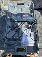 Ryobi hammer drill in open gray plastic case with power cord and manual visible. Drill shows paint marks and signs of use.