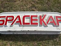 Large white fibreglass sign with raised red letters spelling 'SPACEKAP'. Dirt and grime visible, placed on grass.