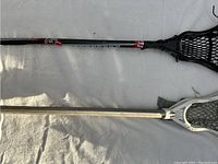 Photo showing two lacrosse sticks, one with black head and net, the other with white head and net lying parallel on a light surface.